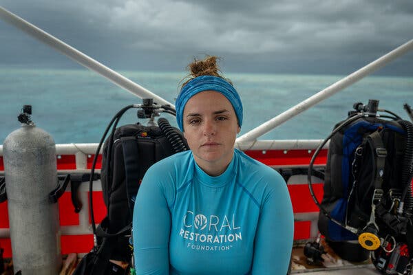 Bailey Thomasson aboard a diving boat. She wears a turquoise shirt and a matching hair band. Her expression is serious. Behind her, two sets of scuba tanks. The sea is calm, but the sky is gray. 